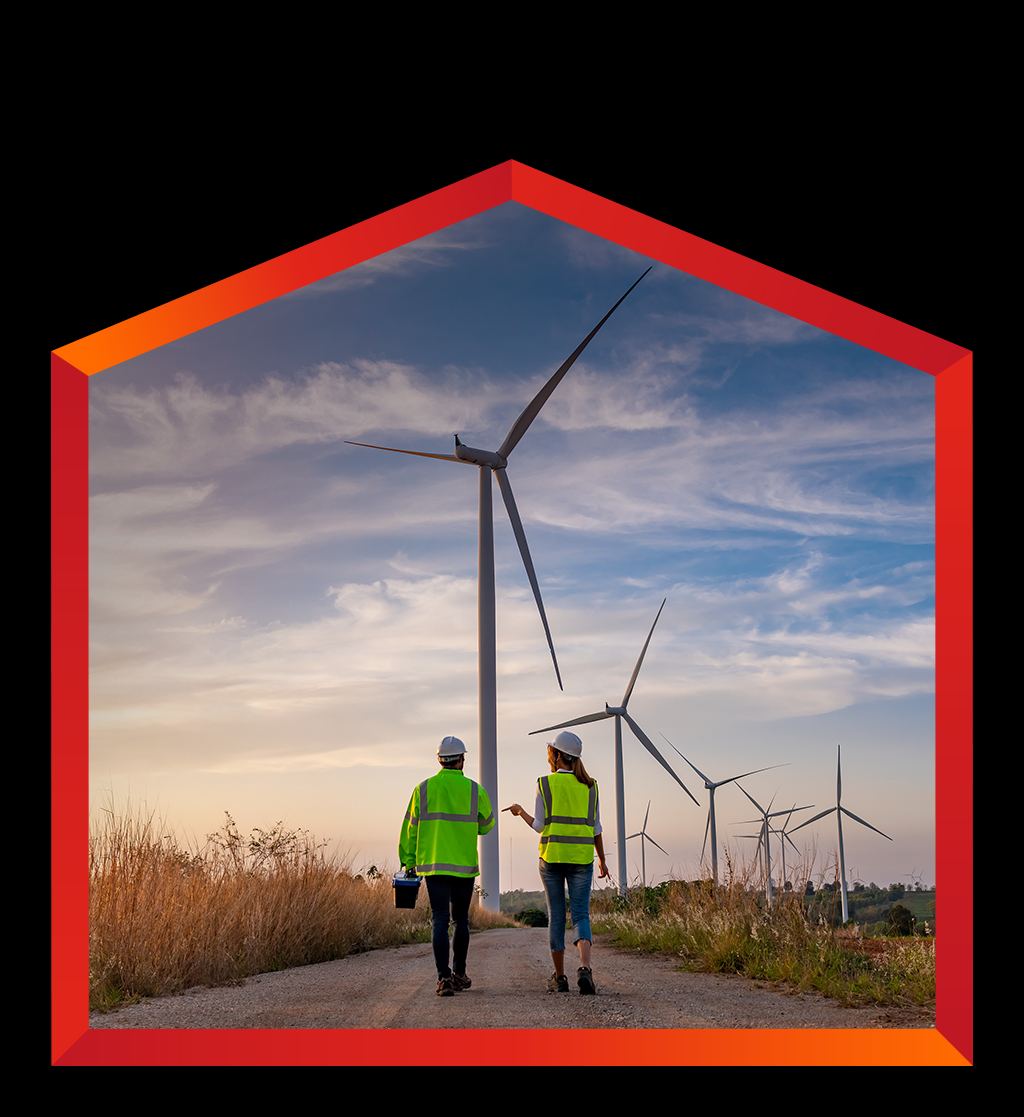Two individuals, seen from behind, walk down a dirt road towards several wind turbines in the distance. Both wear safety vests (one green, one yellow) and hard hats, suggesting they are inspecting a wind farm in Canada.