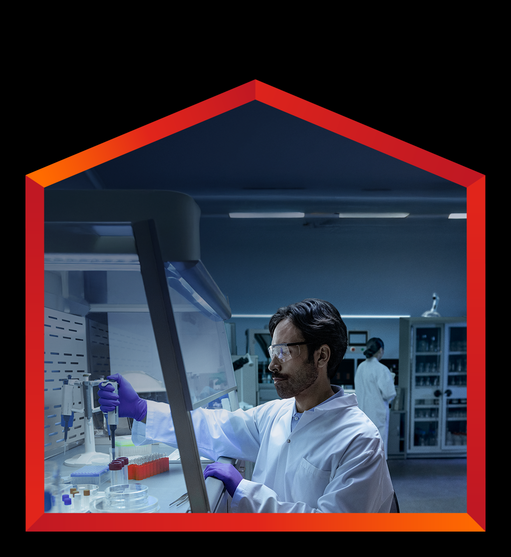 A male scientist in a white lab coat, purple gloves, and safety glasses works inside a fume hood, carefully pipetting liquid into a small beaker in Canada. 