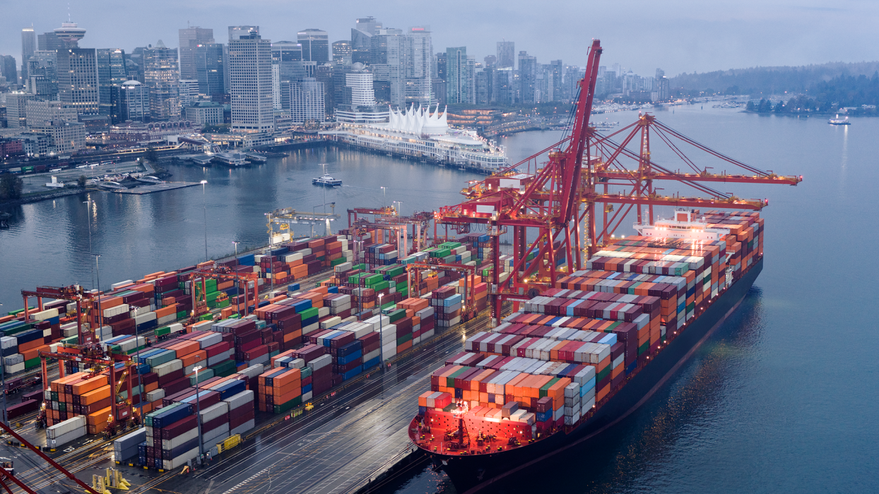 An aerial view shows a massive container ship docked at a port, heavily loaded with thousands of colorful shipping containers. Large red gantry cranes extend over the ship, ready for loading or unloading in Canada.