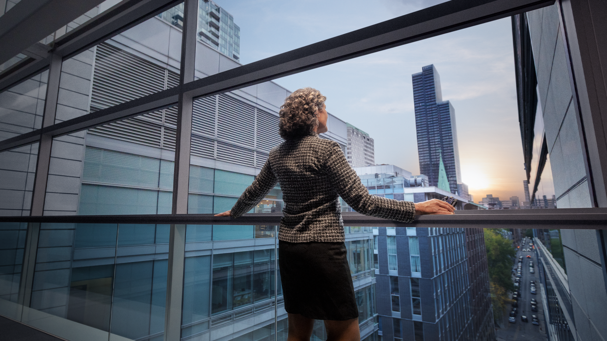 A female is seen from behind, standing and looking out a large, floor-to-ceiling window or balcony. The person is wearing a dark, professional-looking jacket, with their hands resting on a dark horizontal railing. Beyond the window, a modern cityscape is visible with multiple tall, glass-fronted buildings in Canada.
