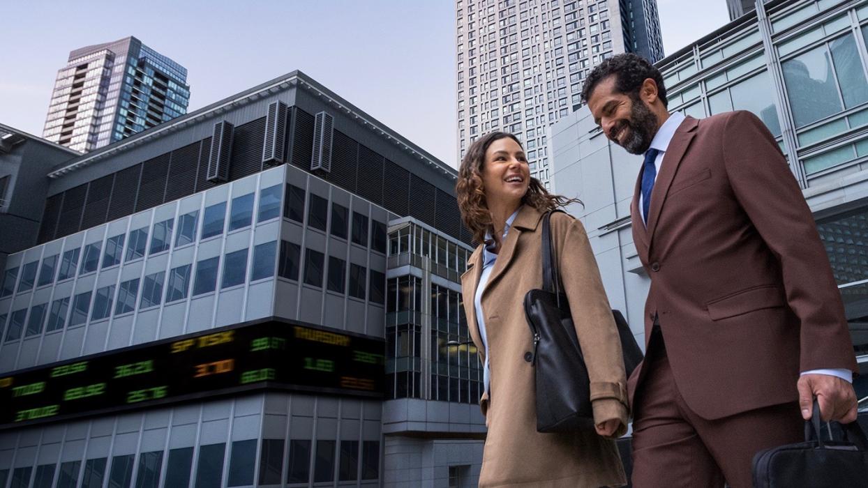 A man in a suit carrying a briefcase and a woman in a beige coat carrying a handbag walk down a wide stone staircase, smiling and looking at each other as if in conversation in Canada.