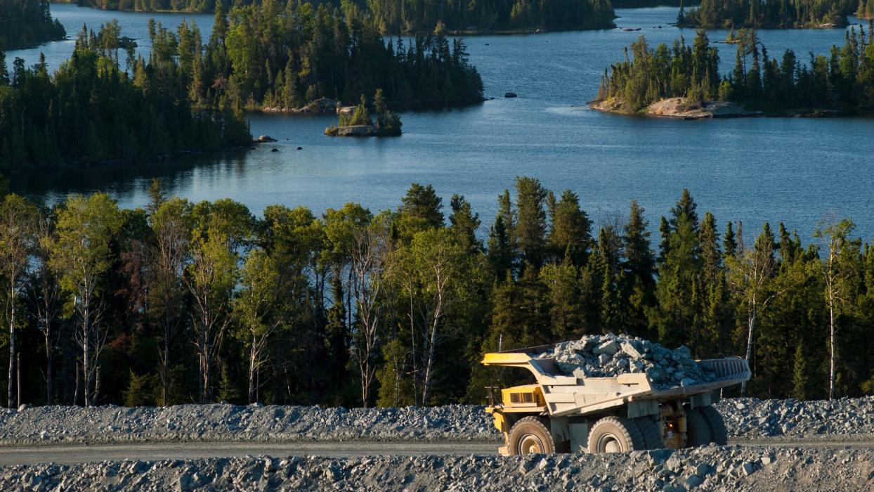 A large, yellow and grey mining dump truck, heavily loaded with rocks, drives on a dirt road in Canada.