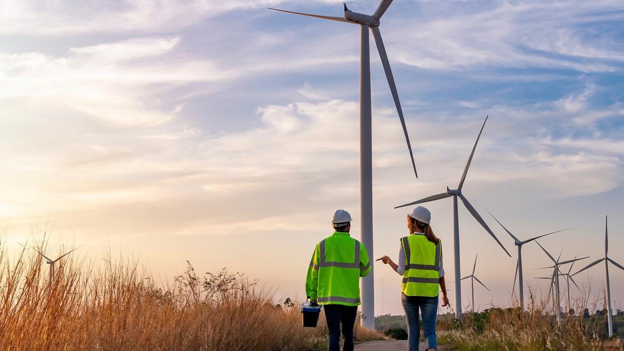 Two individuals, seen from behind, walk down a dirt road towards several wind turbines in the distance. Both wear safety vests (one green, one yellow) and hard hats, suggesting they are inspecting a wind farm in Canada.