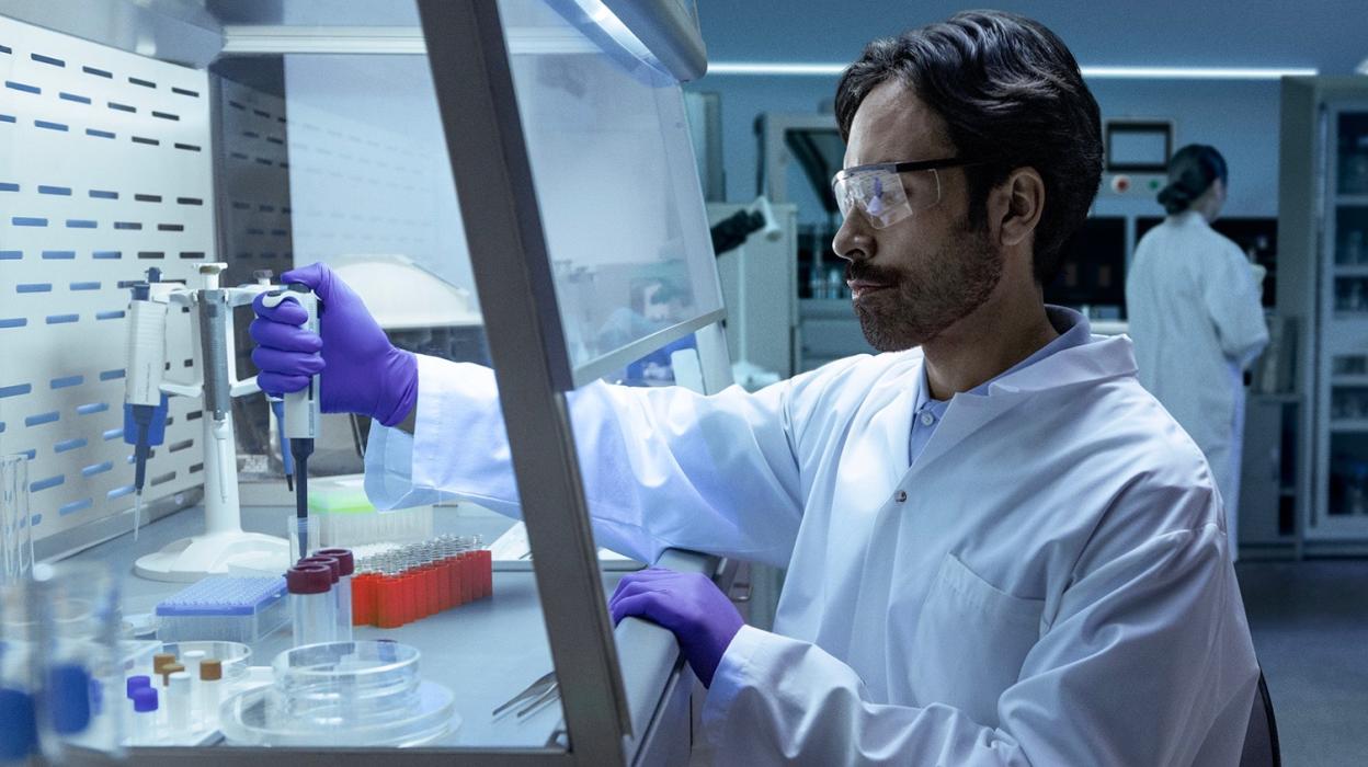 A male scientist in a white lab coat, purple gloves, and safety glasses works inside a fume hood, carefully pipetting liquid into a small beaker in Canada.